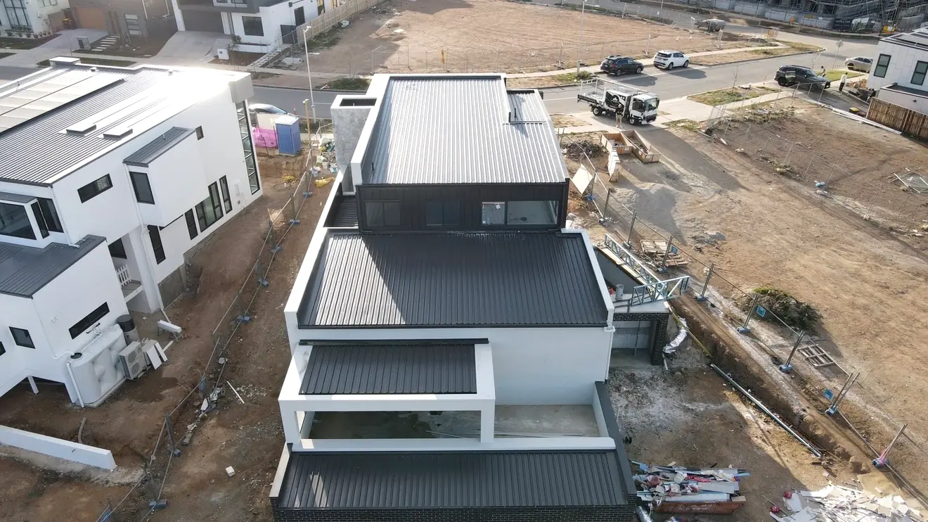 Drone view of a white rendered three level home with dark ribbed metal roofs on each level
