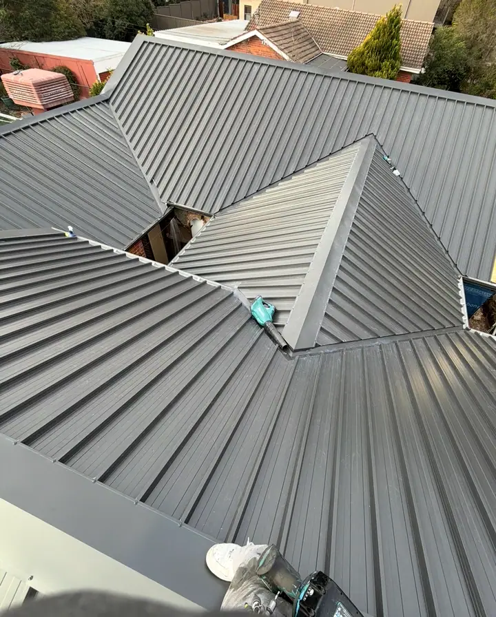 A charcoal ribbed metal roof with hips and a valley, with a blower and drill on the sheets