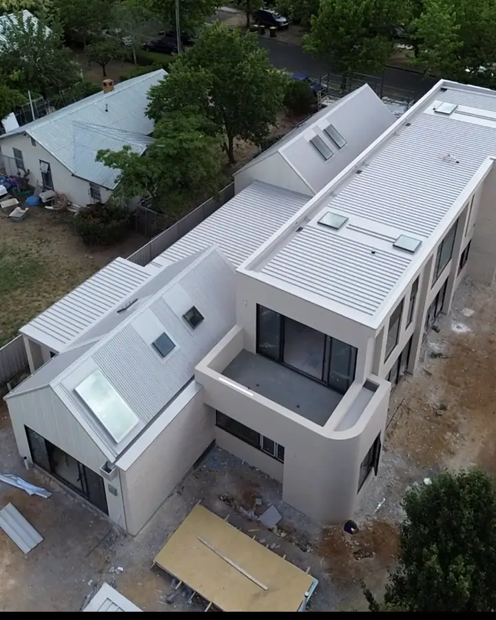 Drone view of a pale two storey home with light metal roofing, vertical wall cladding and several skylights